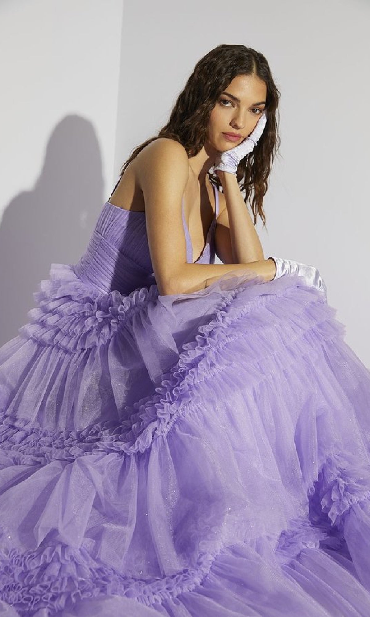 A woman with wavy hair sits pensively in the Lace-Up Long Glitter-Tulle Prom Dress: Eureka 9888 and white gloves, set against a plain light background.